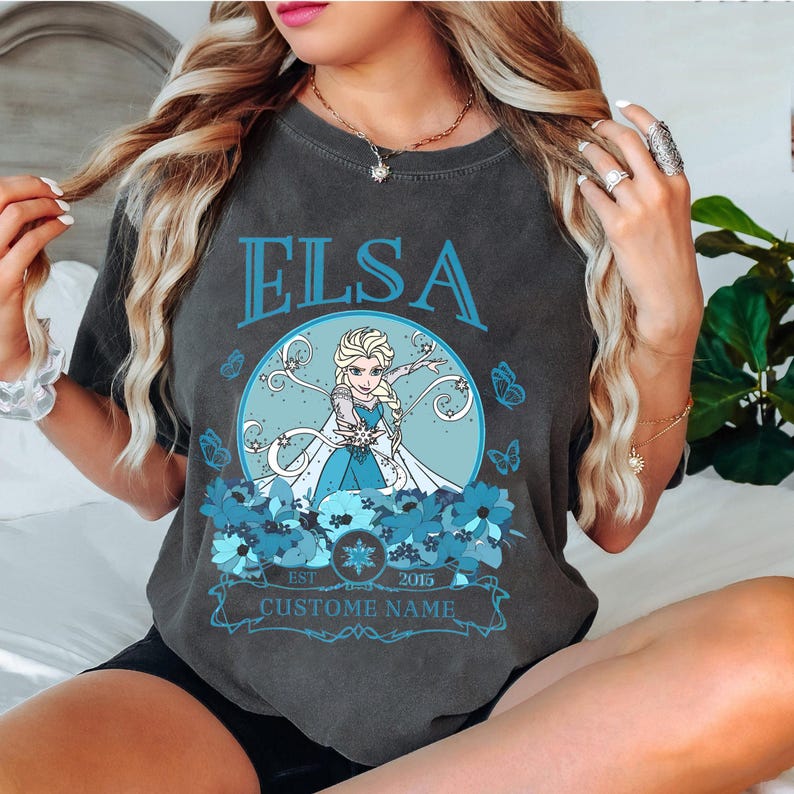 K&ouml;nnte beinhalten: Ein dunkelgraues T-Shirt mit einem Aufdruck von Elsa aus Frozen. Der Aufdruck zeigt Elsa in einem blauen Kleid mit einem wei&szlig;en Umhang, umgeben von blauen Blumen. Der Text "Elsa" ist in Blau &uuml;ber dem Aufdruck gedruckt, und "Est. 2015" und "Custome Name" sind unter dem Aufdruck gedruckt.