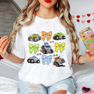 May include: White t-shirt with a colorful design of monster trucks and bows. The trucks are blue, green, and yellow with black wheels. The bows are orange, yellow, light blue, and green.