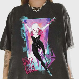 May include: Dark gray t-shirt featuring a graphic of Spider-Gwen in a dynamic pose against a cityscape backdrop. The character is depicted in a black and white suit with pink and white accents. The text "Spider Gwen" is displayed in pink and green.