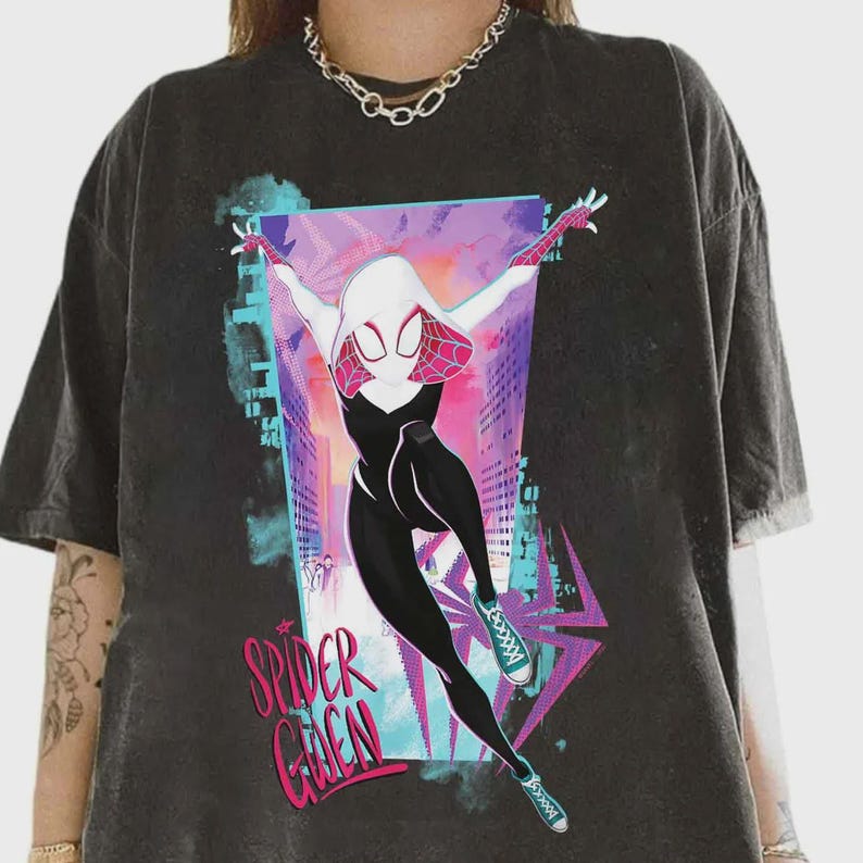 May include: Dark gray t-shirt featuring a graphic of Spider-Gwen in a dynamic pose against a cityscape backdrop. The character is depicted in a black and white suit with pink and white accents. The text "Spider Gwen" is displayed in pink and green.