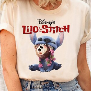Może przedstawiać: Kremowy t-shirt z grafiką z filmu Lilo & Stitch Disneya. Projekt przedstawia Stitcha, niebieskiego kosmitę, trzymającego brązowego misia. Tekst "Disney's Lilo & Stitch" jest wydrukowany na czerwono i czarno nad grafiką.
