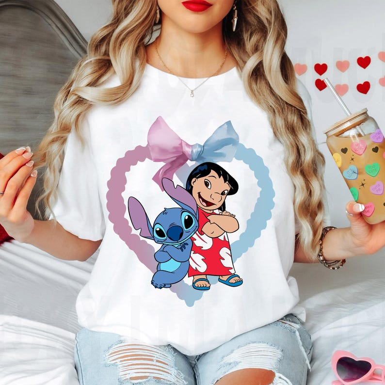 May include: White t-shirt with a blue and purple heart design featuring Stitch and Lilo from Disney's Lilo & Stitch. The heart has a pink and blue bow on top.