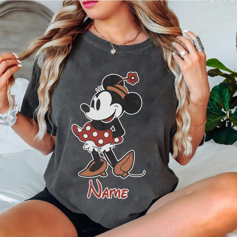 Peut inclure: Un t-shirt gris avec une image de dessin anim&eacute; vintage de Minnie Mouse portant une jupe rouge &agrave; pois et des chaussures rouges. Le texte "NAME" est imprim&eacute; sous l'image.