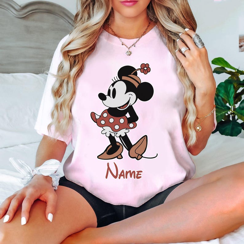 Peut inclure: T-shirt rose avec une image de dessin anim&eacute; de Minnie Mouse portant une jupe &agrave; pois rouges et une fleur dans les cheveux. Le texte "NAME" est imprim&eacute; sous l'image.