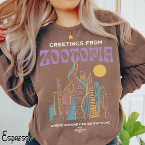 May include: Espresso-colored sweatshirt featuring the text "Greetings From Zootopia" in purple, with a stylized city skyline graphic. The design includes a yellow sun and the phrase "Where anyone can be anything."