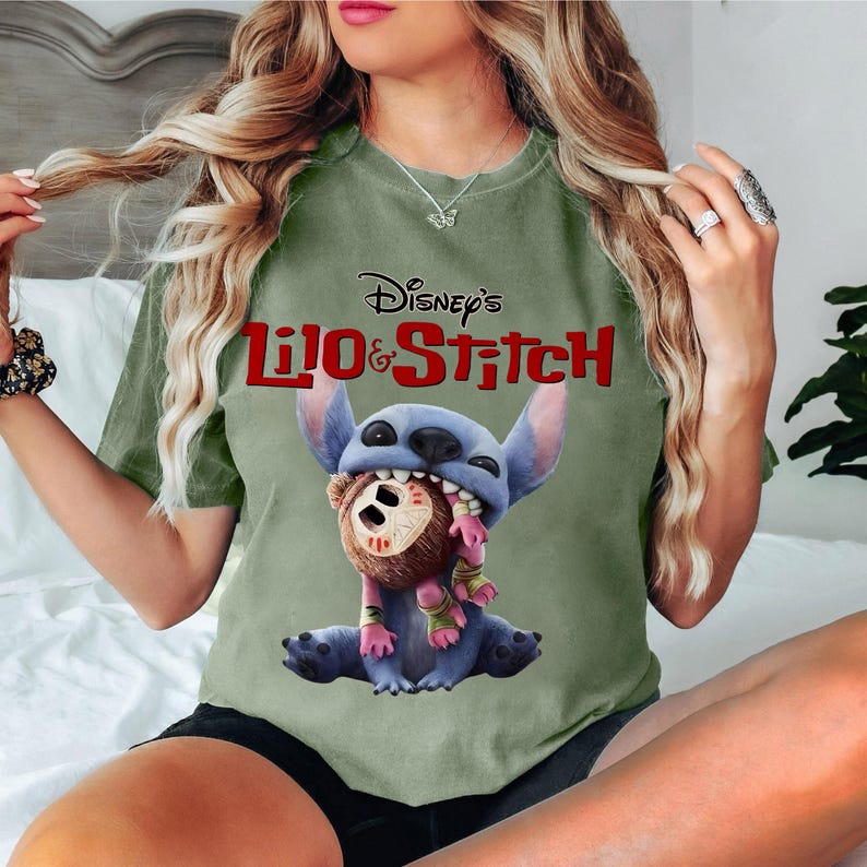 Może przedstawiać: Oliwkowa koszulka z czerwonym logo Disney's Lilo & Stitch. Grafika przedstawia Stitcha trzymającego brązową lalkę z maską. Koszulka ma swobodny styl.