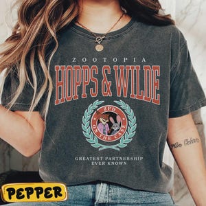 Op de afbeelding: Donkergrijs t-shirt met de tekst "ZOOTOPIA HOPPS & WILDE" in rood en wit. Daaronder een cirkelvormig embleem met "ZPD ON THE CASE" en cartoonfiguren. De zin "GREATEST PARTNERSHIP EVER KNOWN" staat eronder.