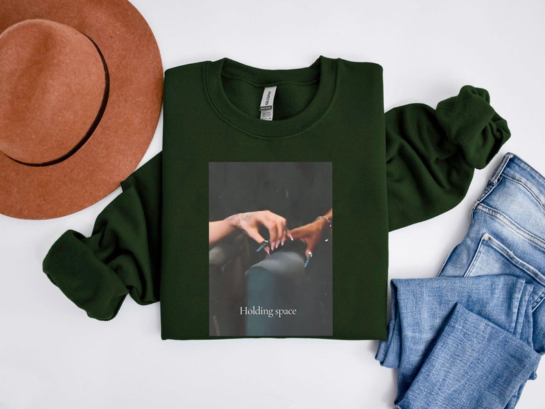 Pu&ograve; includere: Una felpa verde scuro con un'immagine rettangolare di due mani che si tengono. L'immagine ha il testo "Holding space" in basso. Un cappello marrone e jeans blu sono anche nell'immagine.