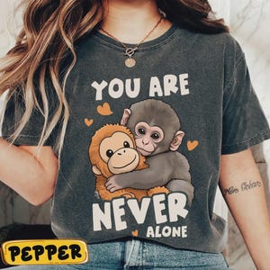 May include: Dark grey t-shirt with a cartoon illustration of two monkeys embracing. The text "YOU ARE NEVER ALONE" is printed above and below the monkeys. Small orange hearts are scattered around the monkeys.