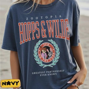 Op de afbeelding: Marineblauwe t-shirt met de tekst "ZOOTOPIA HOPPS & WILDE" in rode en witte letters. Een cirkelvormige afbeelding toont stripfiguren en de woorden "ZPD ON THE CASE". De zin "GREATEST PARTNERSHIP EVER KNOWN" staat eronder gedrukt.