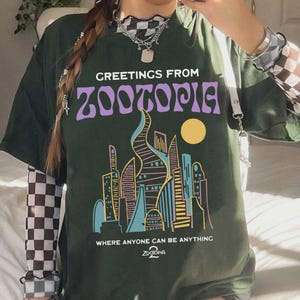 May include: Dark green t-shirt with the text "GREETINGS FROM ZOOTOPIA" in purple. Below the text is a colorful cityscape with a yellow sun. The shirt also has the text "WHERE ANYONE CAN BE ANYTHING" and the Zootopia logo.