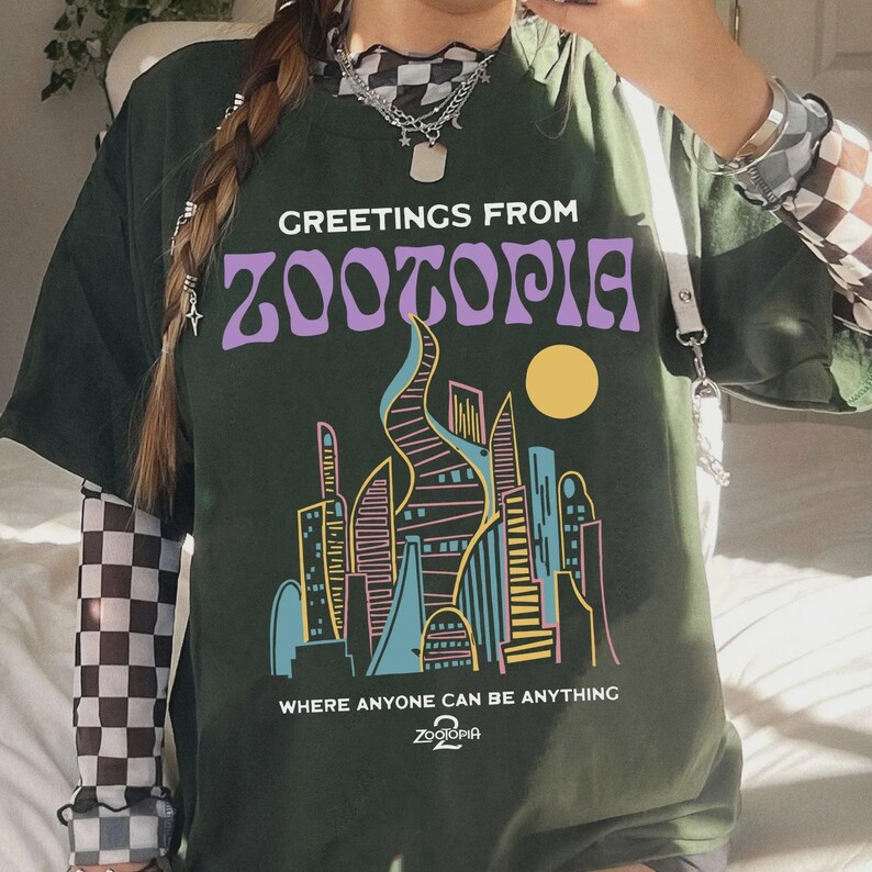 May include: Dark green t-shirt with the text "GREETINGS FROM ZOOTOPIA" in purple. Below the text is a colorful cityscape with a yellow sun. The shirt also has the text "WHERE ANYONE CAN BE ANYTHING" and the Zootopia logo.