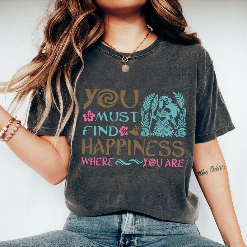 May include: A dark gray t-shirt with a graphic print that says "You must find happiness where you are". The graphic features a stylized image of a woman with a flower in her hair and a spear in her hand.