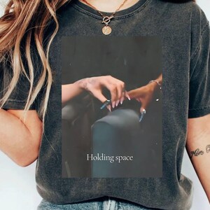 Pu&ograve; includere: T-shirt grigio scuro con una grafica rettangolare raffigurante due mani che si toccano, con lunghe unghie blu. Il testo "Holding space" &egrave; stampato sotto. La maglietta ha un girocollo e maniche corte.