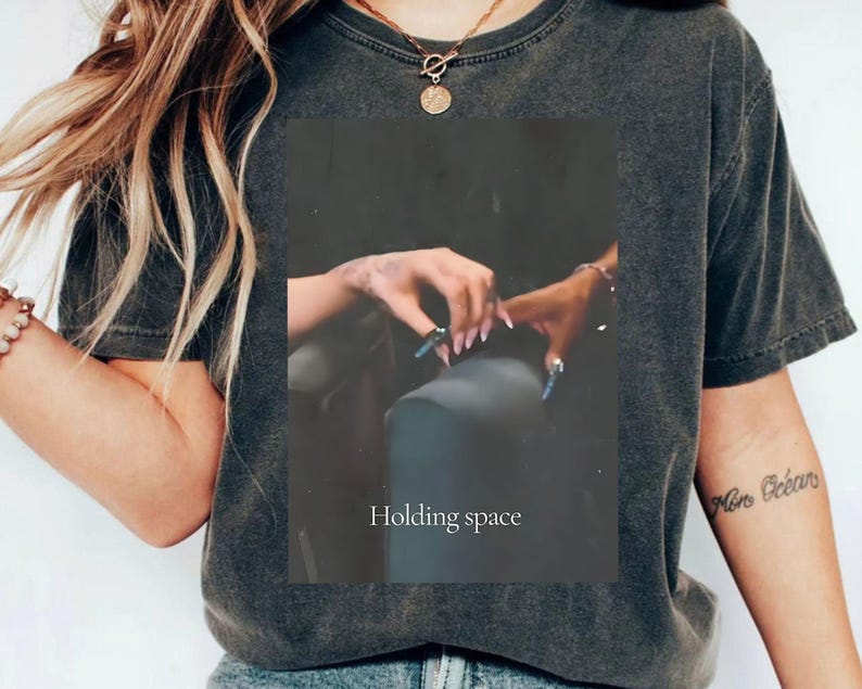 Pu&ograve; includere: T-shirt grigio scuro con una grafica rettangolare raffigurante due mani che si toccano, con lunghe unghie blu. Il testo "Holding space" &egrave; stampato sotto. La maglietta ha un girocollo e maniche corte.