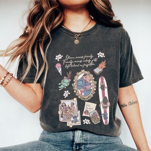 Puede incluir: Camiseta gris oscuro con un collage de ilustraciones de dibujos animados y el texto "Ohana significa familia. Familia significa que nadie se queda atr&aacute;s ni se olvida." El dise&ntilde;o incluye un helado, una criatura verde y una imagen enmarcada de una pareja.