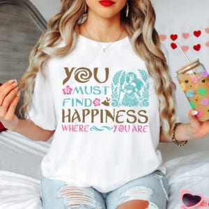 May include: White t-shirt with a brown and blue graphic design featuring a cartoon character and the text "You must find happiness where you are."