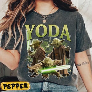May include: Dark grey t-shirt with the word "YODA" in green block letters above three illustrations of Yoda with green lightsabers. The illustrations are set against a background of green lightning.
