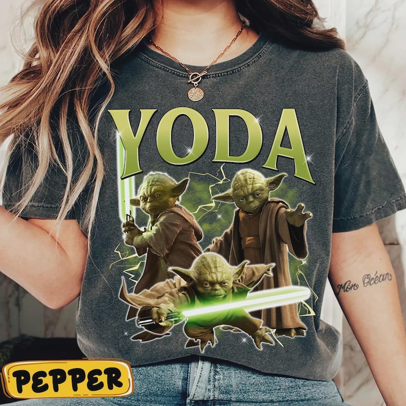 May include: Dark grey t-shirt with the word "YODA" in green block letters above three illustrations of Yoda with green lightsabers. The illustrations are set against a background of green lightning.