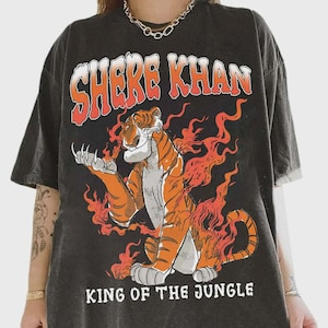 Puede incluir: Camiseta gris oscuro con una ilustración de tigre y llamas naranjas. El texto "SHERE KHAN" está encima del tigre, y "KING OF THE JUNGLE" debajo. El tigre es naranja y blanco.