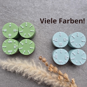 Könnte beinhalten: Zwei Sätze von vier runden, hellgrünen und hellblauen Objekten mit weißen Markierungen und dem Text "Viele Farben!" in Braun. Die Objekte haben Zahlen und Symbole. Ein Zweig trockenes Gras befindet sich im Vordergrund.