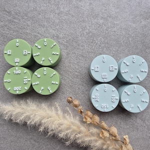 Könnte beinhalten: Sechs runde, grüne und blaue Kunststoffknöpfe mit aufgedruckten Zahlen und Symbolen. Die Knöpfe sind in zwei Reihen zu je drei angeordnet.