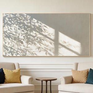 May include: A large rectangular wall art piece featuring a neutral beige background with a prominent shadow pattern of tree leaves cast across the left side. The artwork is framed in a thin light wood border and hangs centered above two armchairs.