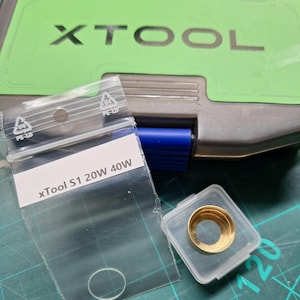 Puede incluir: Una bolsa de plástico transparente con una etiqueta blanca que dice "xTool S1 20W 40W". Un pequeño anillo de metal dorado está en un recipiente de plástico transparente.