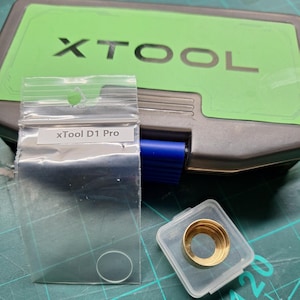 Puede incluir: Una bolsa de plástico transparente con una etiqueta blanca que dice "xTool D1 Pro" y un pequeño objeto redondo transparente en su interior. Un pequeño recipiente de plástico transparente con un anillo de metal dorado en su interior. El recipiente está sobre una superficie de rejilla verde y negra.