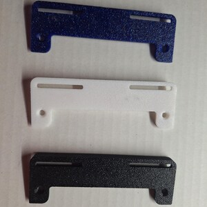 May include: Three textured brackets in blue, white, and black. Each bracket has two rectangular slots and two circular holes. The brackets have a unique shape with a curved section on one end.