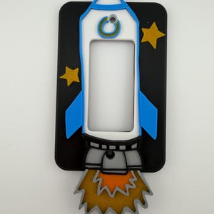 May include: A decorative light switch cover featuring a cartoon rocket ship design. The rocket is white and blue, with a black background and gold stars. The rocket's exhaust is orange and gold.