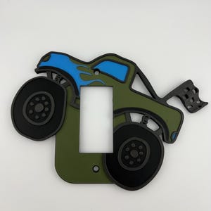 May include: A decorative light switch cover in the shape of a monster truck. The cover is olive green with black wheels and a blue flame design on the cab. A checkered flag is attached to the back of the truck.