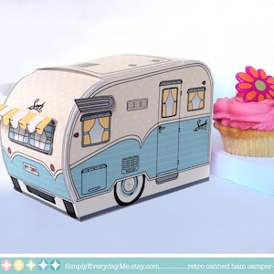 May include: A blue and white camper trailer shaped box with a yellow and white striped awning. The box is decorated with a flower and the word "Sweet". The box is sitting on a white surface next to a pink cupcake with a pink flower on top.