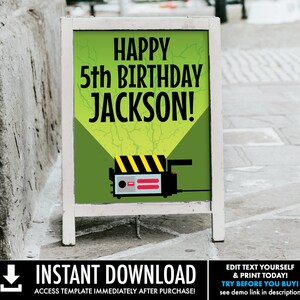 May include: A green and yellow sign with the text "Happy 5th Birthday Jackson!" and a cartoon image of a ghost trap. The sign is on a white easel.