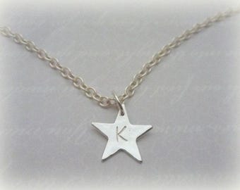 May include: A silver-colored necklace featuring a star pendant. The star has a single letter "K" engraved on it and hangs from a delicate chain. The necklace is displayed against a soft, blurred background.