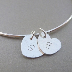 May include: A silver bangle bracelet with two silver heart charms. The charms are engraved with the letters "G" and "E".