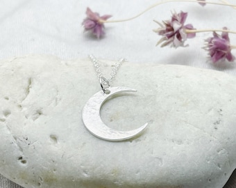 May include: A silver crescent moon pendant necklace. The pendant has a textured surface and hangs from a delicate chain. The necklace is displayed on a white stone with dried purple flowers in the background.