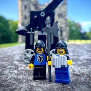 May include: Two toy knights stand in front of a toy castle and catapult. The knight on the left is wearing blue armor with a black eagle on his chest. The knight on the right is wearing blue armor and holding a silver cup. Both knights have black helmets and yellow faces.