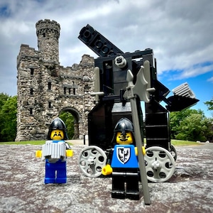 May include: Two black Lego knights stand in front of a black Lego catapult. The catapult is on a stone surface in front of a stone castle with an arched doorway. The knights are wearing black helmets and armour, and one is holding a black spear.
