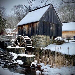 Puede incluir: Una rueda de agua de madera con un edificio de piedra detrás. La rueda está cubierta de nieve y el edificio está parcialmente cubierto de nieve. Hay un arroyo de agua que fluye frente a la rueda.