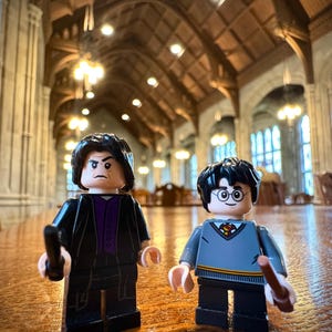 Pode incluir: Duas minifiguras Lego, uma com uma túnica preta e a outra com um suéter cinza, estão em pé sobre uma mesa de madeira. A minifigura com a túnica tem uma varinha mágica na mão. A minifigura com o suéter tem uma varinha mágica na mão. As minifiguras estão em uma grande sala abobadada com um teto de madeira.