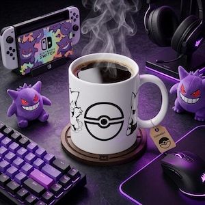 Puede incluir: Una taza de cerámica blanca con un gráfico negro de una Poké Ball y personajes Gengar, llena de líquido oscuro humeante. La taza descansa sobre un posavasos de madera circular, acompañada por dos peluches de Gengar morados y equipo de juego.