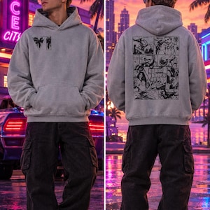 Puede incluir: Una sudadera con capucha gris claro que presenta un pequeño gráfico negro de palmeras y el número romano VI en el pecho. La parte posterior muestra un gran estampado negro estilo cómic con el texto Grand Theft Auto Vice City.