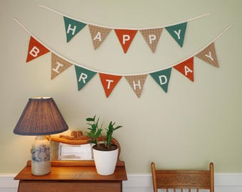 Könnte beinhalten: Zwei festliche Geburtstagsbanner hängen an einer Wand. Das obere Banner zeigt "HAPPY" mit grünen, orangefarbenen und gemusterten Wimpeln. Das untere Banner zeigt "BIRTHDAY" mit ähnlichen Farben. Eine Lampe und eine Pflanze stehen auf einem Holztisch.