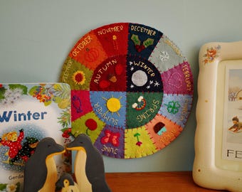 Puede incluir: Una colorida rueda de fieltro que representa las estaciones, con detalles bordados y texto. Un libro infantil titulado "Winter" y figuras de pingüinos de madera están en primer plano. Una imagen enmarcada con el texto "FEBRUARY" está a la derecha.