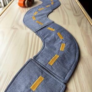 May include: A blue fabric road with yellow stitched lane markings. A small orange wooden toy car sits nearby. The road is made of three connected pieces, creating a winding path. The road is placed on a light wood surface.