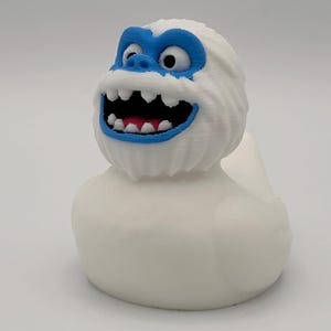 Op de afbeelding: Een witte badeend met een yeti-ontwerp. De eend heeft een witte body, een blauw gezicht en mondcontour en een rode tong. Het gezicht van de eend heeft een vriendelijke uitdrukking met een wijd open mond en zichtbare tanden.