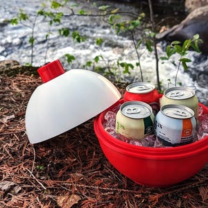 Puede incluir: Una nevera roja y blanca con forma de boya de pesca, abierta para revelar cuatro latas de bebida sobre hielo. La nevera está sobre un lecho de agujas de pino marrones cerca de un arroyo.