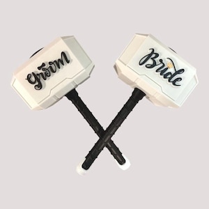 May include: Two white toy hammers with black handles are crossed. One hammer has the word "Groom" in black script, and the other has "Bride" with a small crown. The hammers are likely wedding decorations.