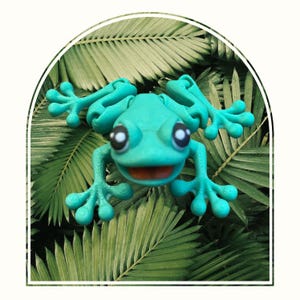 Op de afbeelding: Een felgroen kikkerfiguur met een brede glimlach en grote ogen, geposeerd op een bed van groene palmbladeren.